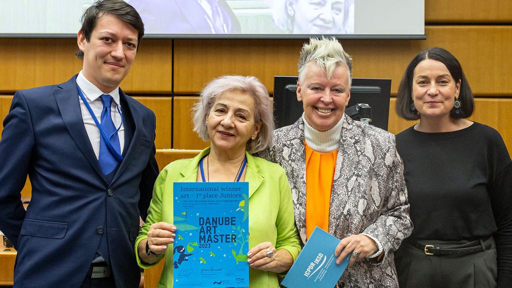 4 persons in a large meeting room in from of a large screen, the second from left a woman holding a blue certificate