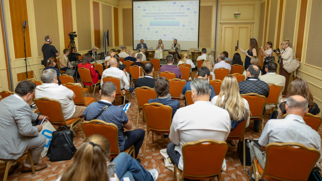 A crowded room with journalists and participants adn a few people on the stage behind a screen 
