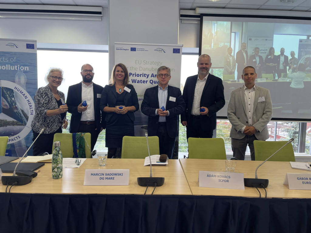 © EUSDR PA4 Six individuals are facing the camera - with a larege screen behind their backs as well as roll ups. They are staning behind green chairs in front of a large desk
