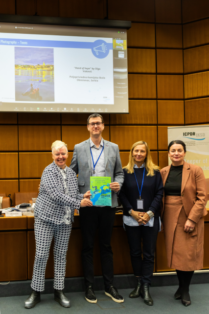 Three women stand with one man holding up colourful certificate. 