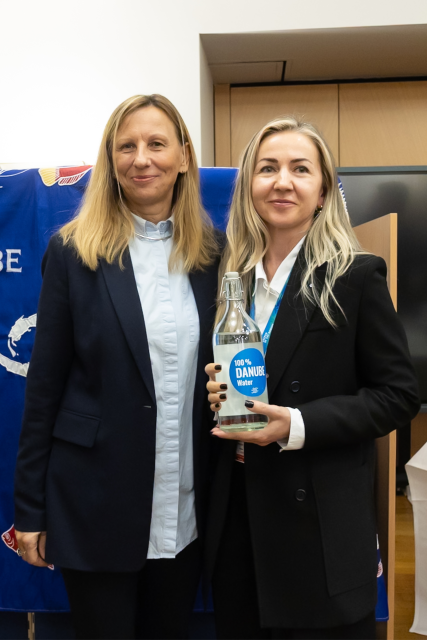 Two women stand next to each other, one holding a glass bottle with a blue sticker and filled with water.
