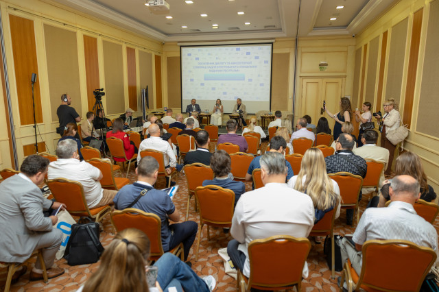 A crowded room with journalists and participants adn a few people on the stage behind a screen 