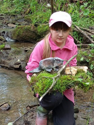 Young girl squats in river with a beaver art piece. 